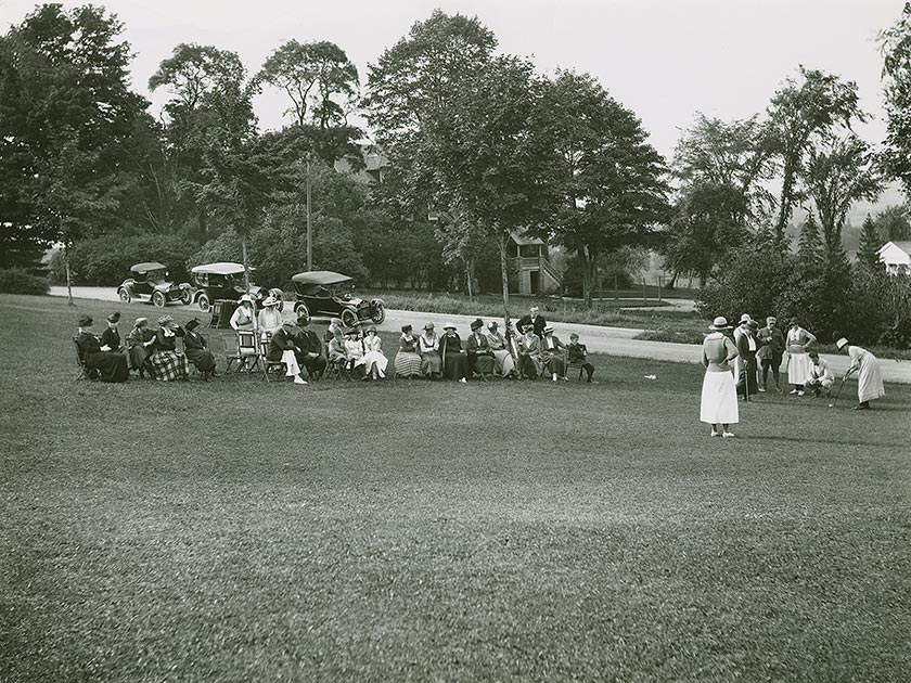 A woman in a long dress prepares to put in a group of golfers, observed by a group in lawn chairs