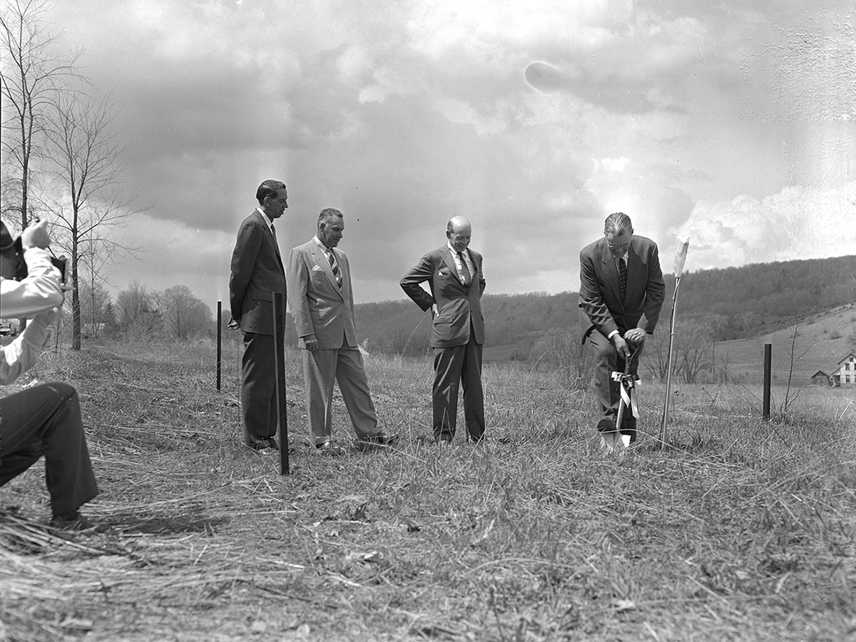 Three men observe as a fourth breaks ground with a shovel. A photographer snaps a photo from the left.