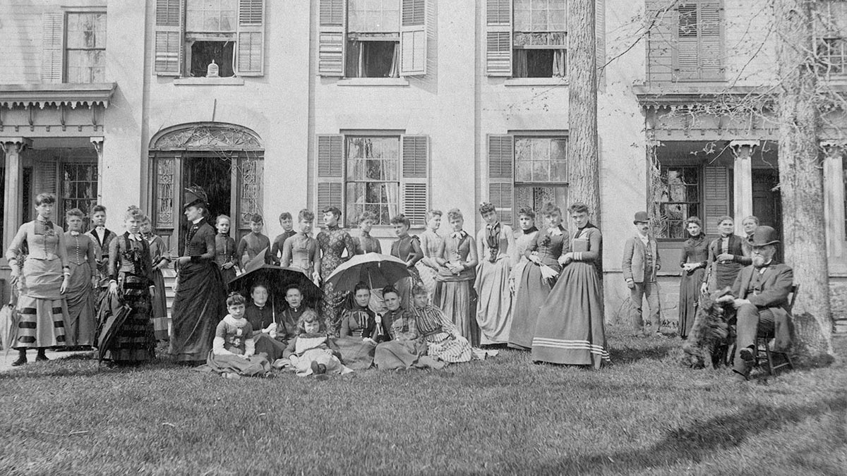Women in full-length dresses and skirts on a building's front lawn with two men in top hats