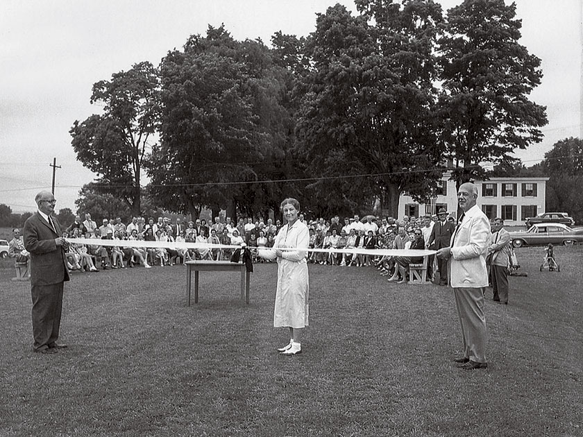 Two men hold a ribbon while a woman cuts it, and a crowd watches. The clubhouse is visible in the background