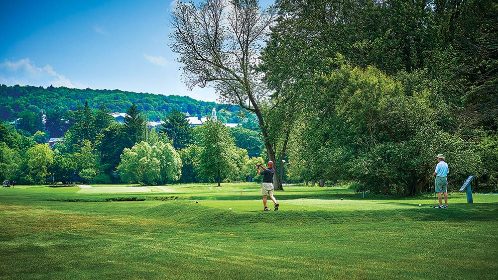 Golfer drives from the tee with the Colgate campus on a hillside in the background